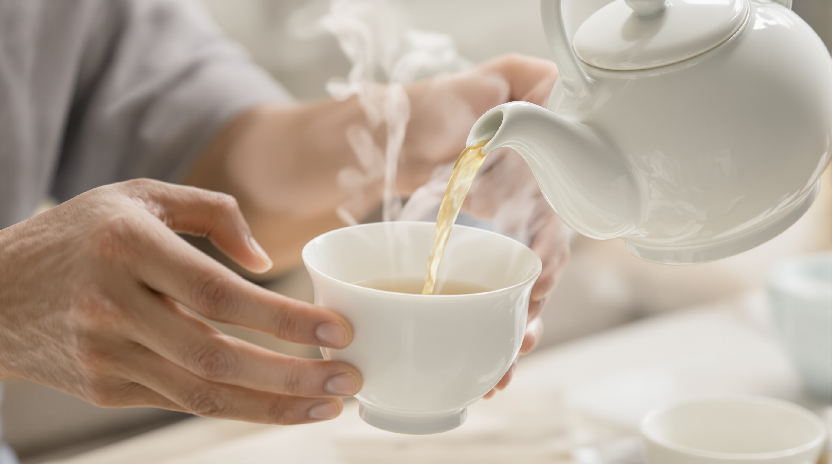 Close-up of hands pouring tea from an ergonomic Chinese porcelain set with curved cups and steam