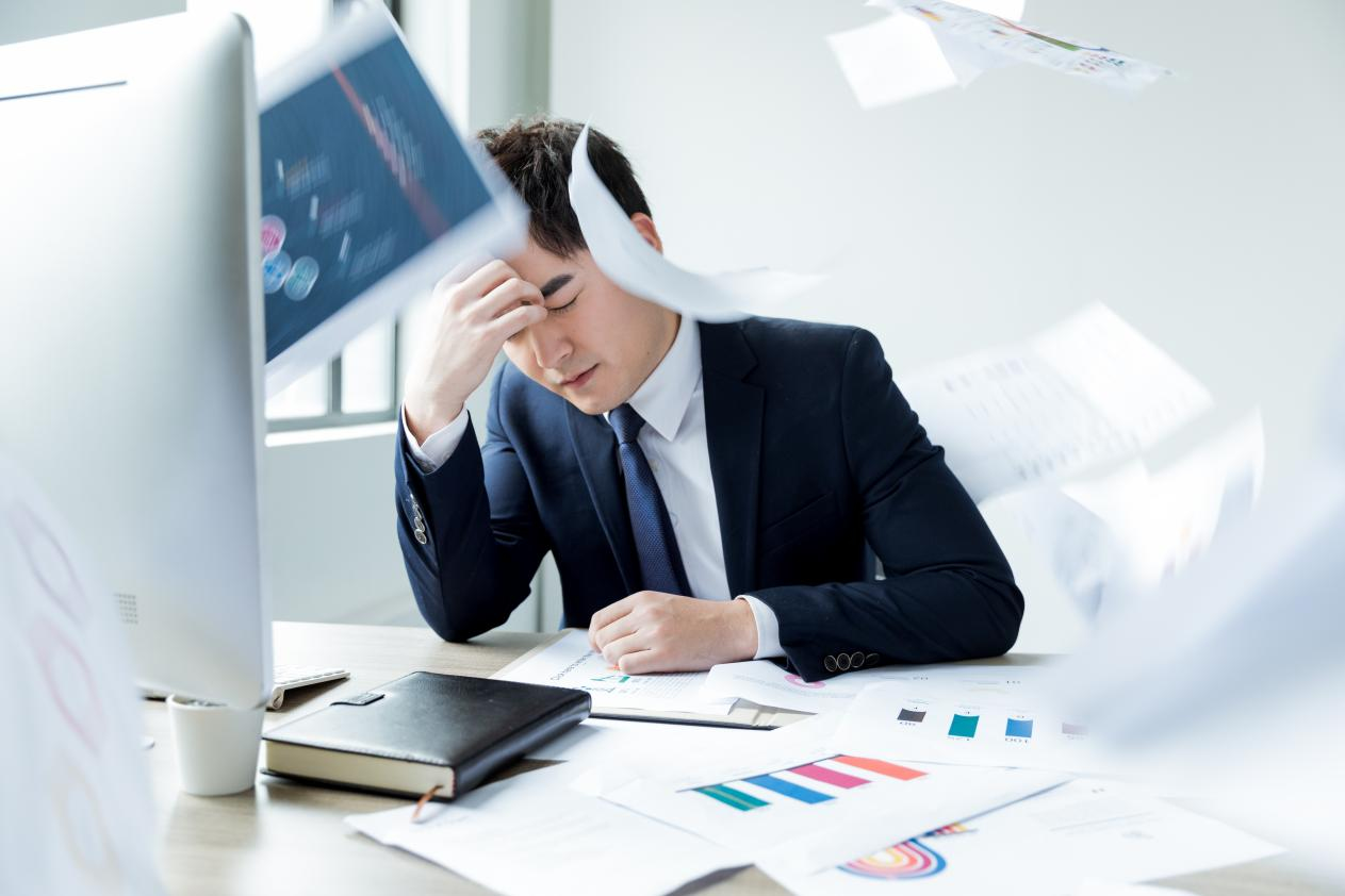An overwhelmed male hotel procurement manager in a suit, looking stressed and exhausted with scattered paperwork, charts, and a computer at his desk, illustrating complex supply chain challenges.