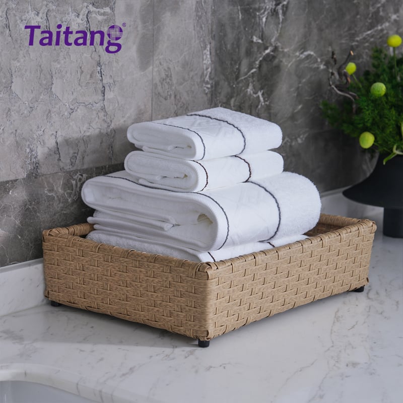 A set of three neatly folded white towels in small, medium, and large sizes, stacked inside a rectangular woven basket sitting on a white marble counterto.jpg