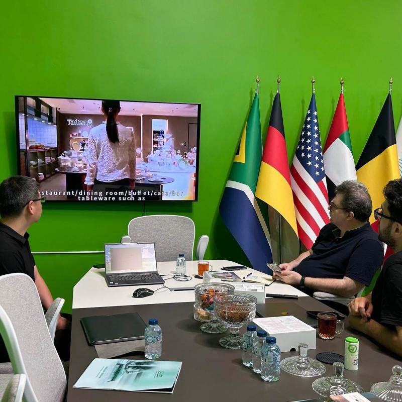 International business clients and Taitang representatives sitting at a conference table with national flags in the background, reviewing promotional videos during the Dubai exhibition.jpg