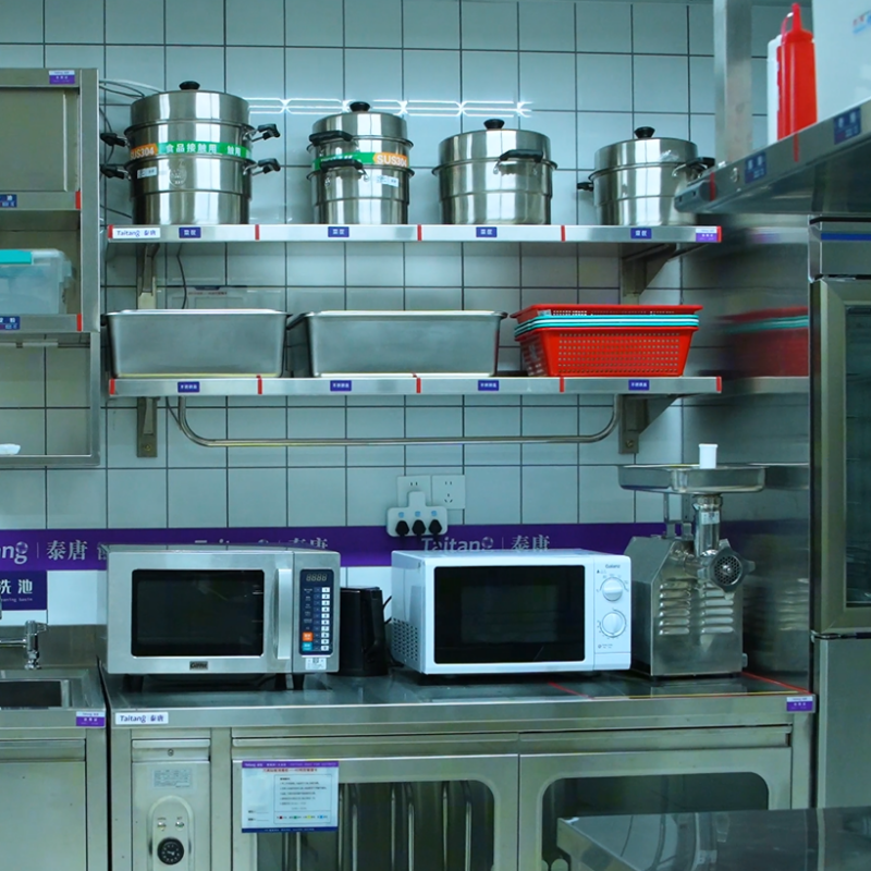 Commercial hotel food preparation area equipped with stainless steel prep counters, commercial microwaves, and wall-mounted storage shelves..png