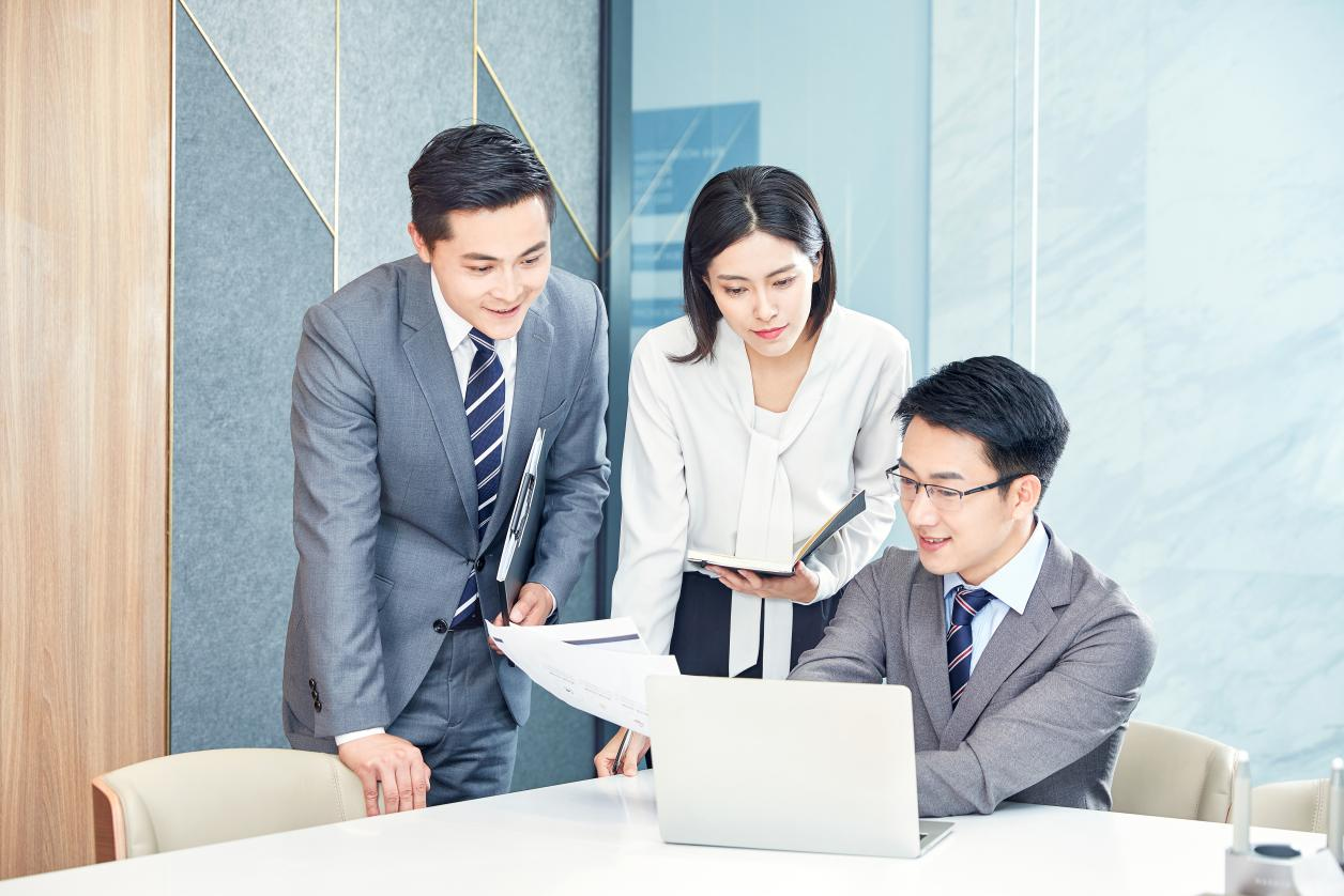 A diverse team of three hotel supply logistics professionals, including a woman holding a notepad, looking at a laptop and documents together during a meeting to coordinate production scheduling for a new hotel project.