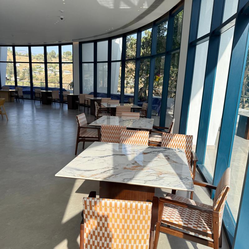 Sunlit resort restaurant with floor-to-ceiling window walls, square stone-top tables, and woven dining armchairs.jpg