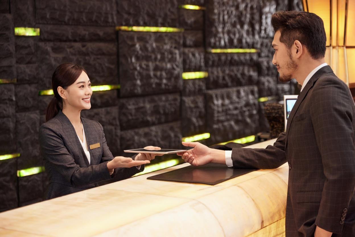 A professional hotel receptionist handing a tablet to a male guest at a modern, high-end marble check-in counter, illustrating detail-oriented hospitality service.