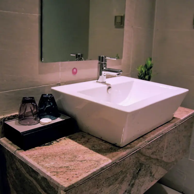 Hotel bathroom washbasin vanity setup featuring a custom black tray with drinking glasses and room amenities.webp