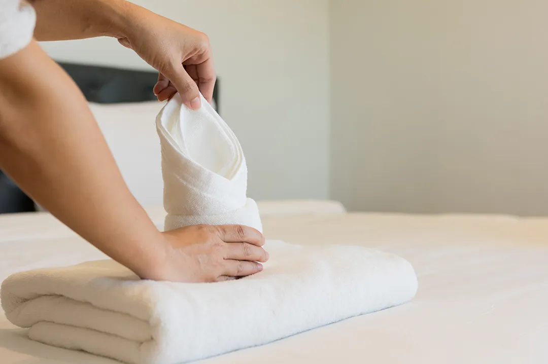 Close-up of hands neatly folding a crisp white guest towel for display on a hotel bed, emphasizing quality guestroom service.