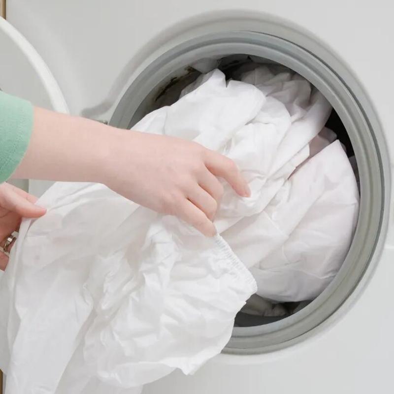 A hand loading white bed sheets into a washing machine, illustrating high-temperature industrial laundering for hotel linens.jpg