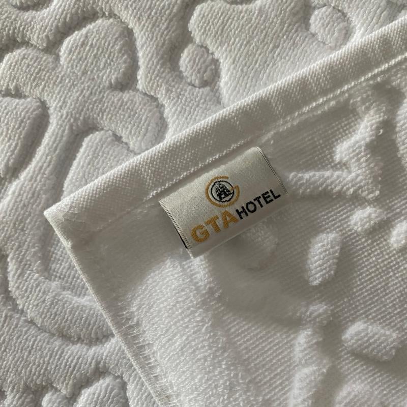 Textured white jacquard hotel bath mat corner displaying a stitched GTA HOTEL woven brand label.JPG