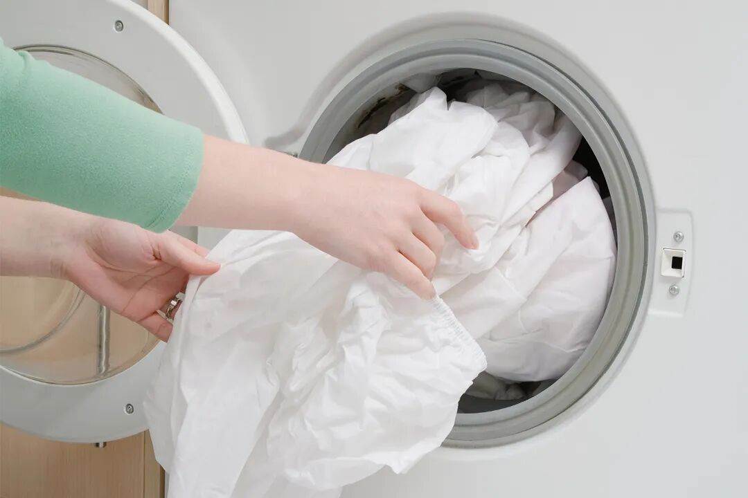 A close-up of white hotel linens being loaded into a professional washing machine, highlighting the durability and bleach-resistance of TAITANG premium fabrics.