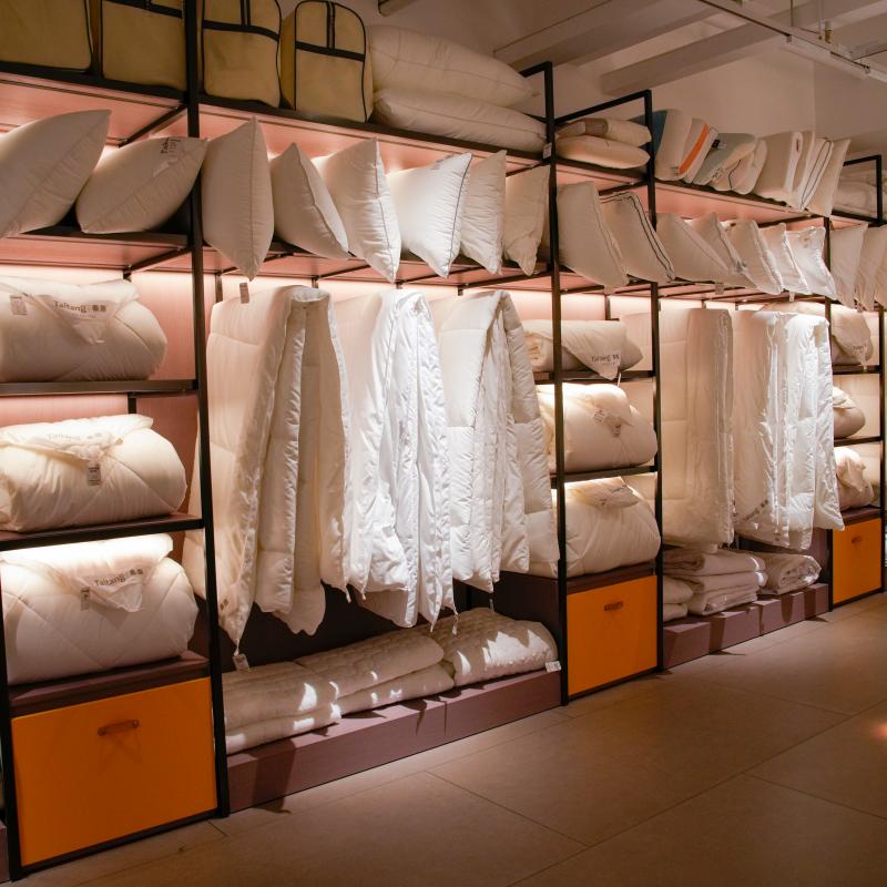 Taitang commercial hotel bedding showroom display featuring bulk wholesale pillows, hanging premium duvets, and comforters on illuminated modern shelving for hospitality procurement managers.JPG