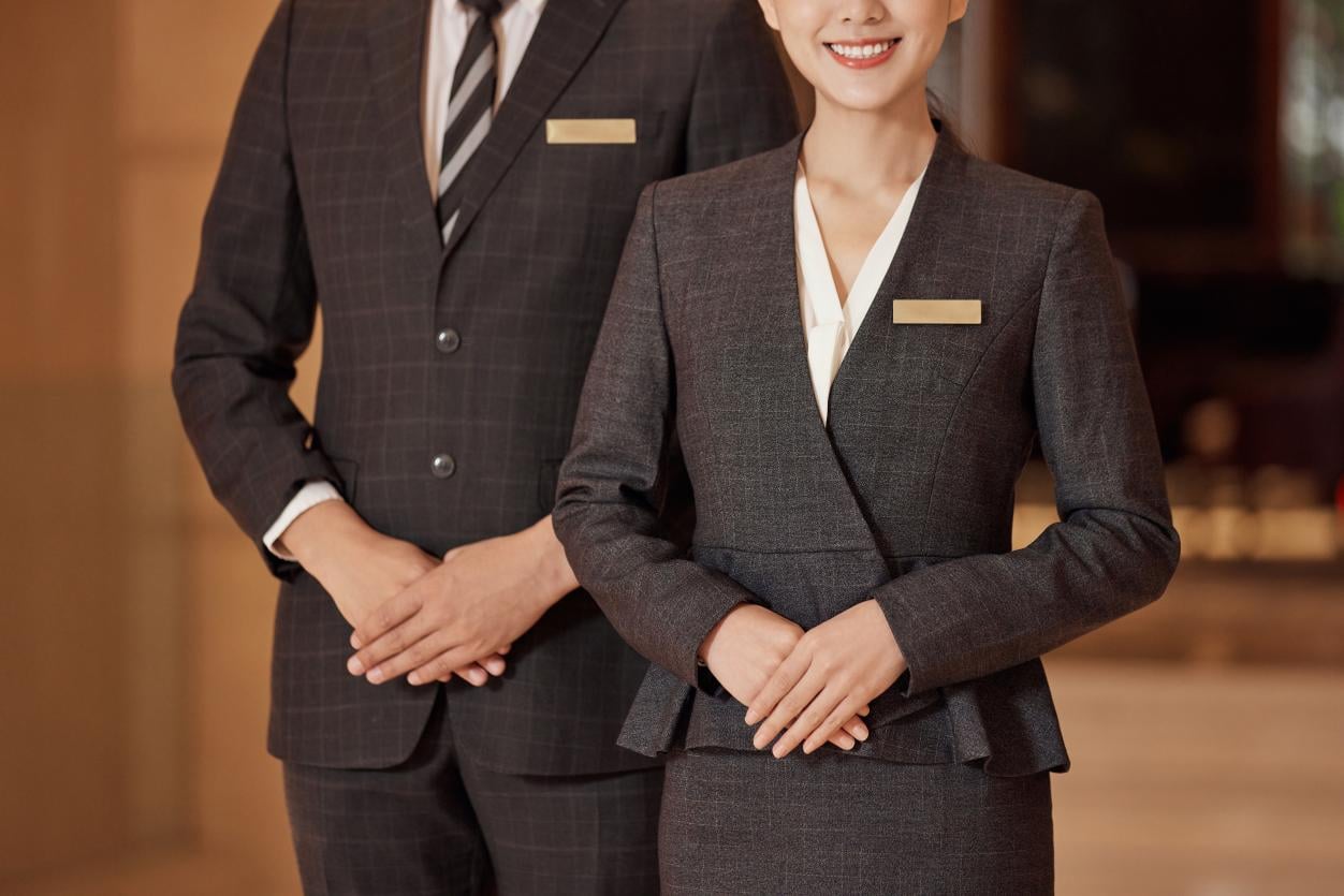 A close-up of a professional male and female hotel staff member in elegant grey uniforms with gold name tags, symbolizing the successful and timely launch of a hotel project supported by Taitang.