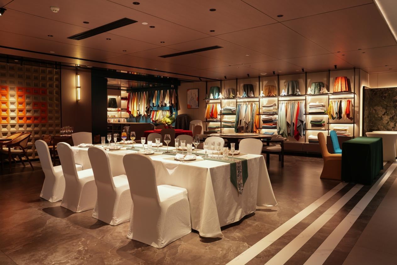 A luxury hotel banquet showroom featuring a long dining table set with white tablecloths, chair covers, and fine glassware, with extensive wall displays of colorful textile samples and restaurant linens in the background.