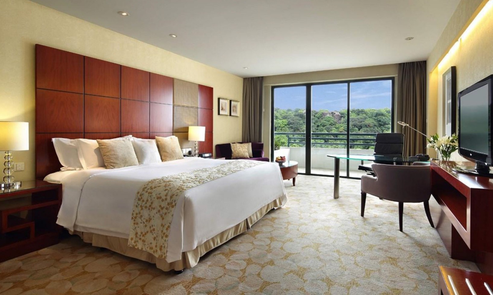 A detailed interior view of a spacious Hilton Guangzhou Jade guest room, equipped with premium hotel room supplies and hotel bedding from Taitang, a leading bed linen company. The room features a king-size bed with crisp white linens, a plush wooden headboard, a well-appointed office area with a glass desk and television, and a balcony with a scenic mountain view.