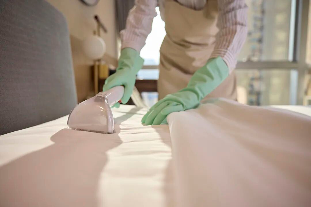 A hotel housekeeping staff member using a professional steam iron on white bedding to ensure a smooth, premium finish and impeccable guest presentation.