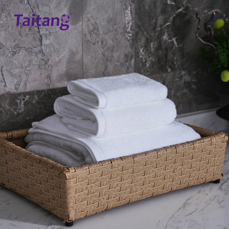 An angled close-up of a neat stack of white luxury towels inside a woven storage basket, highlighting their plush thickness and flat-woven twill borders.jpg