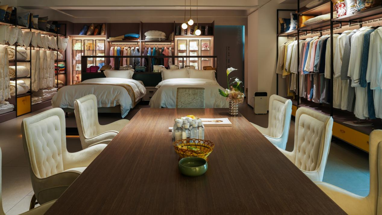 A professional hotel procurement consultation room featuring a large wooden meeting table surrounded by luxury white tufted chairs, with displays of hotel linens, pillows, and bathrobes in the background.