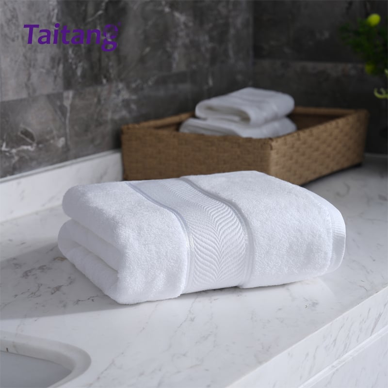 A single plush white bath towel with a wide, decorative shiny satin border, neatly folded and resting on a bright marble vanity countertop.jpg