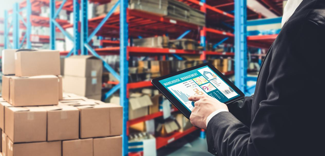 A professional in a suit using a tablet to monitor a digital warehouse management system in a large hotel supply distribution center filled with organized cardboard boxes.