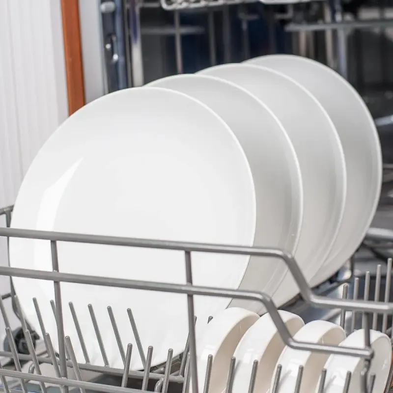 Vitrified commercial white porcelain plates loaded vertically in a dishwasher rack to prevent chipping and glaze damage.webp