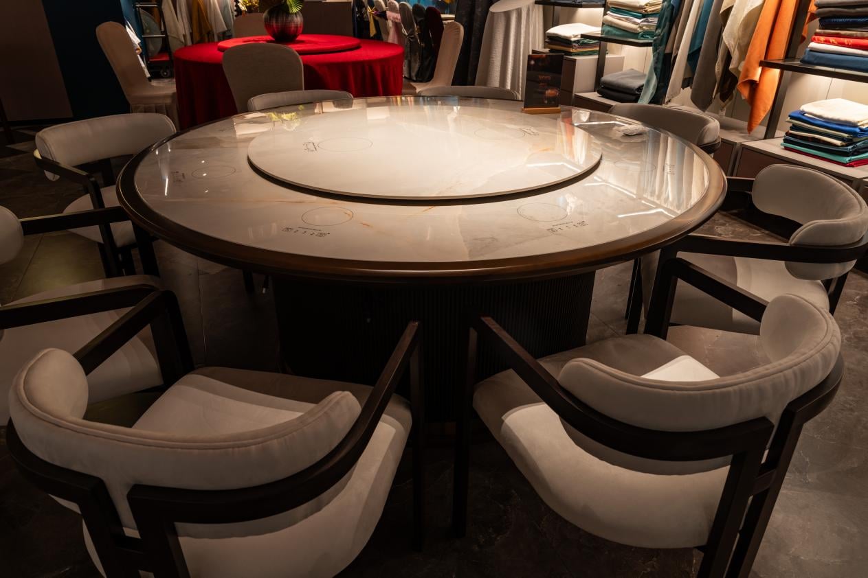 A close-up of a modern, large round dining table with a smooth marble countertop and a built-in Lazy Susan, surrounded by dark wood chairs with white upholstered cushions in a sophisticated dining setting.