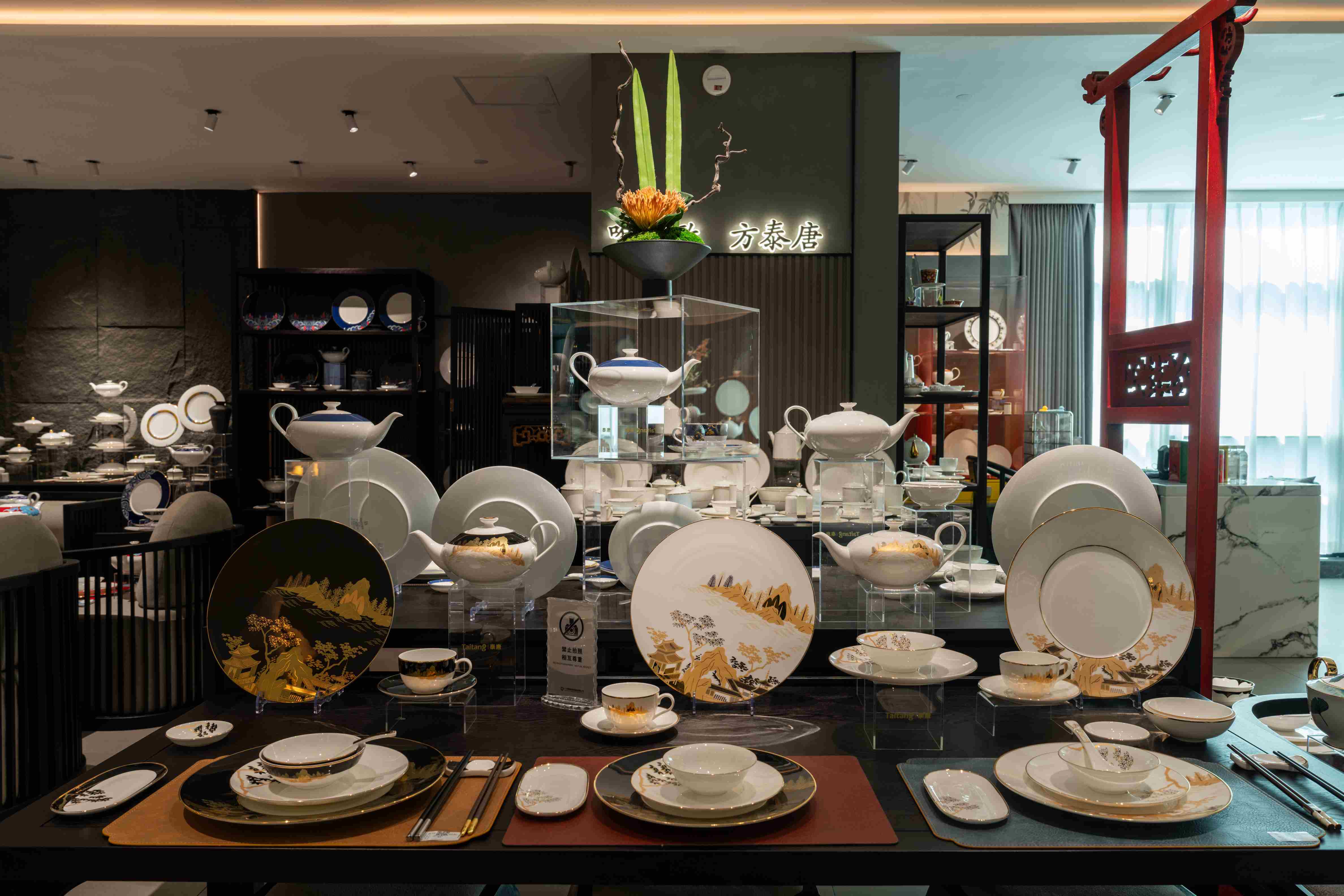 Aluxurious display table in a hotel with high-end dinnerware, including various plates with intricate gold and landscape designs, teapots, and cups.