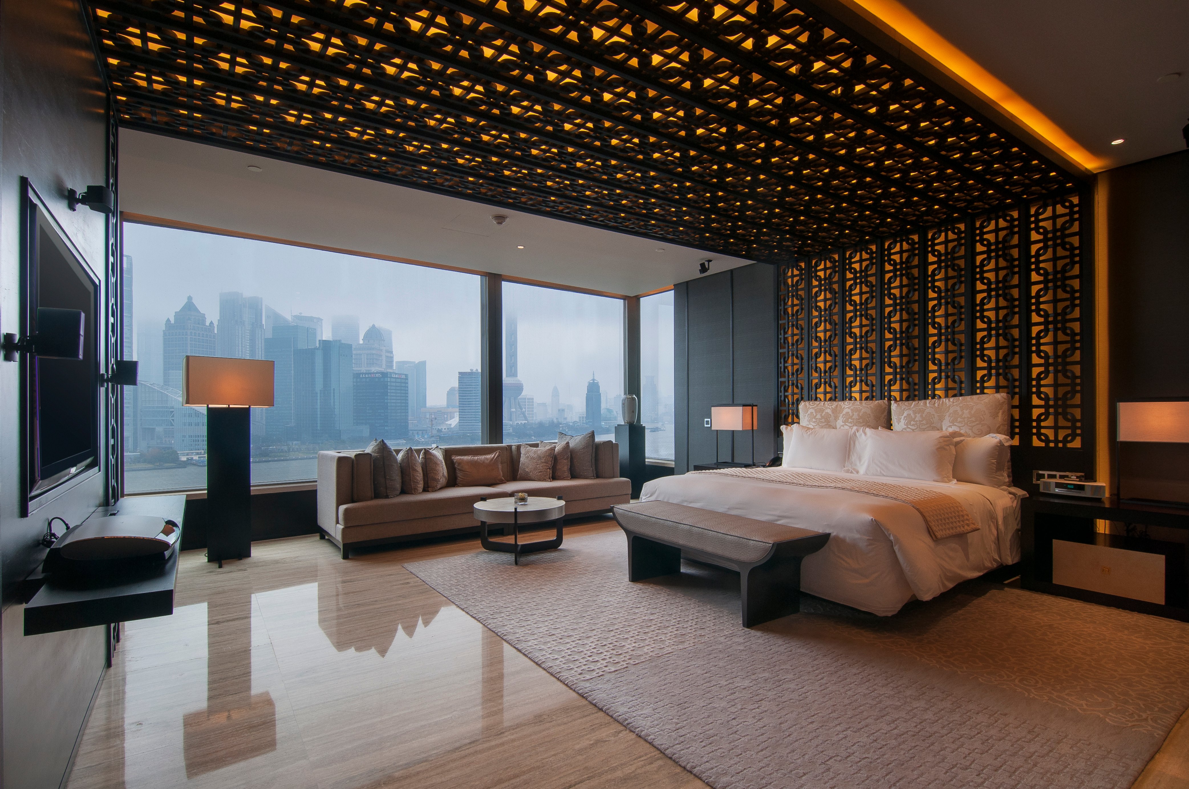 A modern luxury hotel bedroom with a panoramic city skyline view through large floor-to-ceiling windows, featuring a plush bed, a contemporary sofa, and intricate black lattice decorative panels on the ceiling and walls.