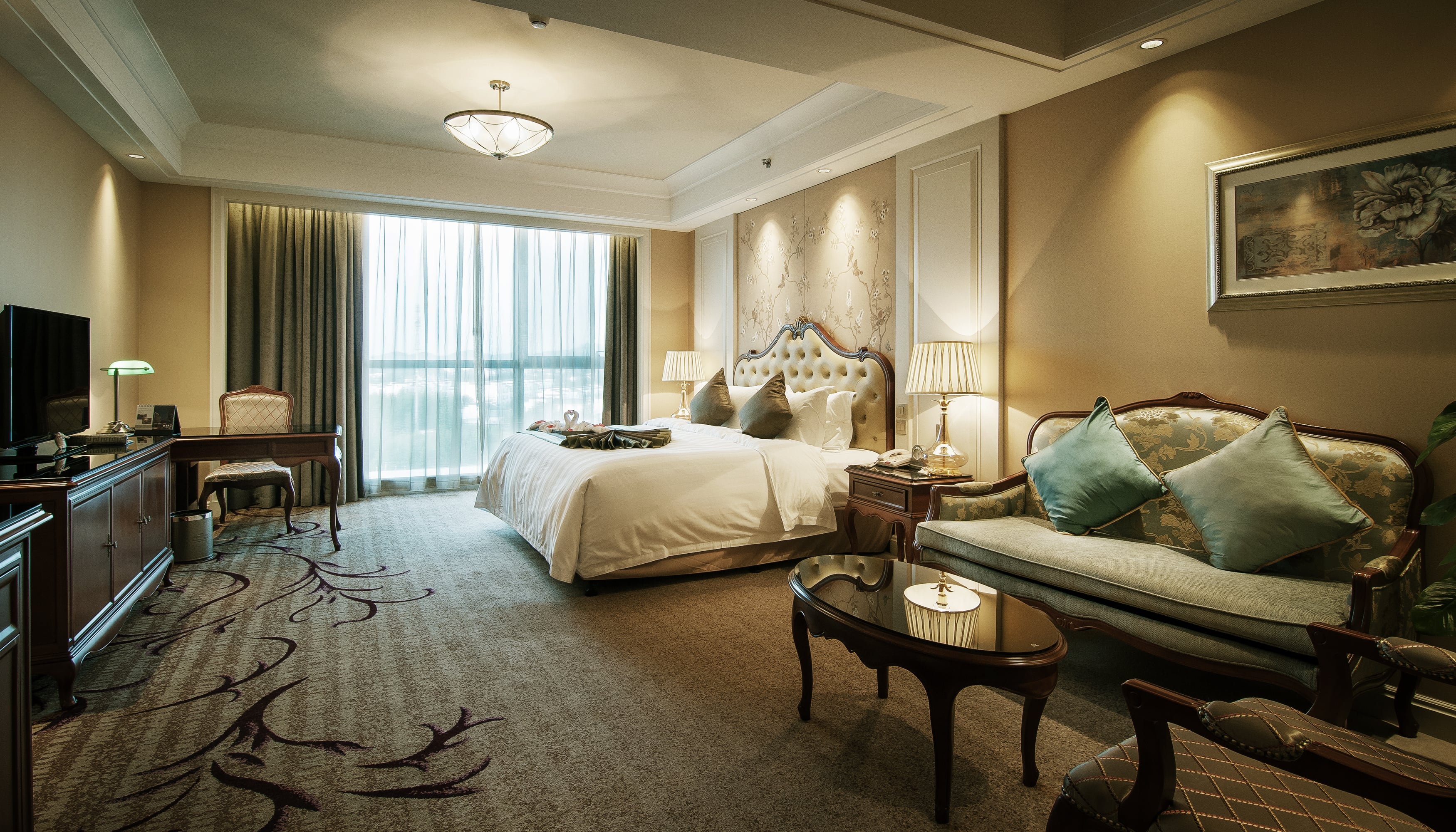 A luxurious hotel suite with a classic interior design, featuring a large bed with a tufted headboard, an elegant green sofa with decorative pillows, a dark wood desk, and a patterned carpet under warm, ambient lighting.