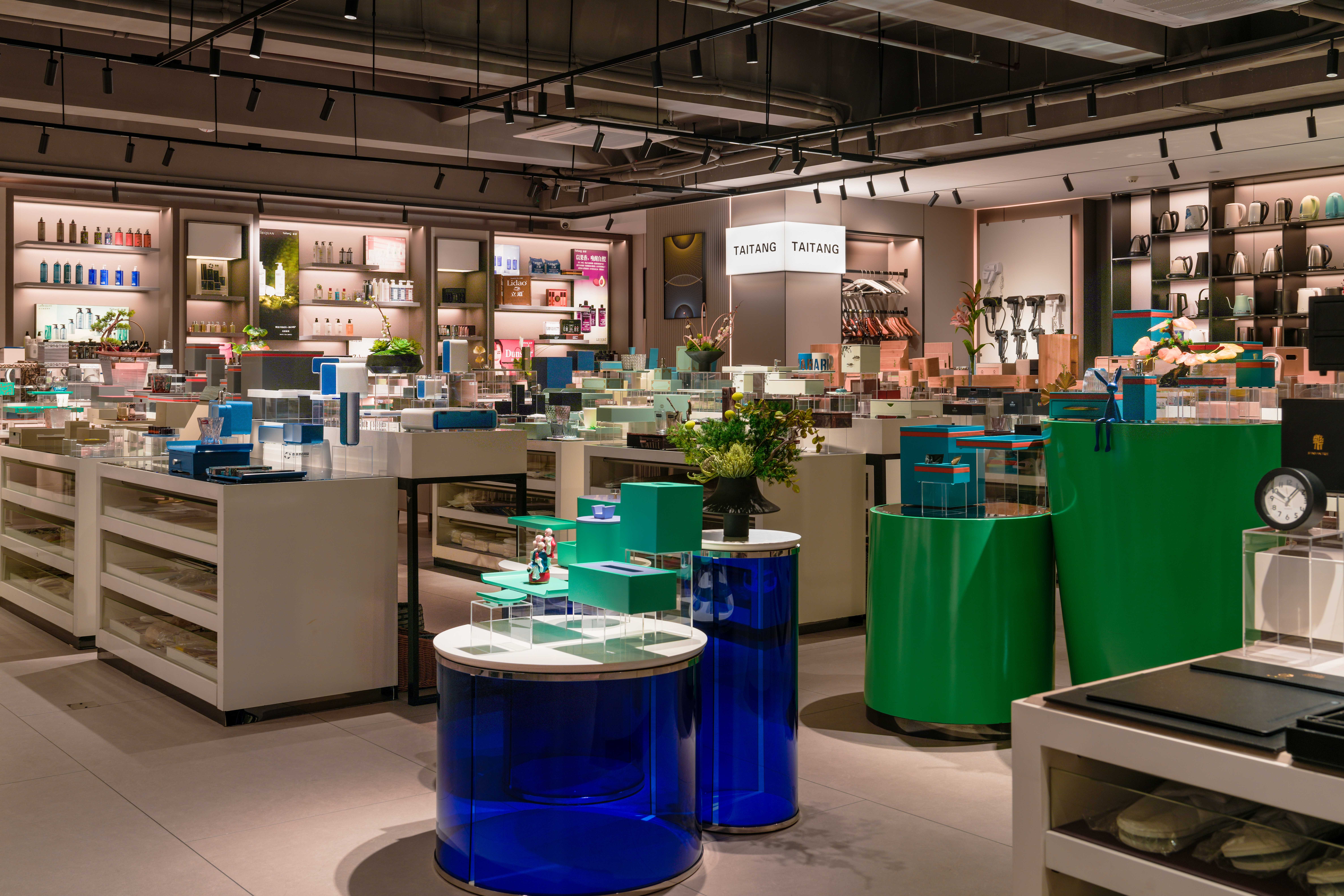 An elegant and brightly lit bathroom amenities and spa products display within a Taitang showroom, featuring multiple curated counters, a striking blue glass display table, and green shelving with luxurious personal care items and plush towels, designed to inspire hotel procurement managers.