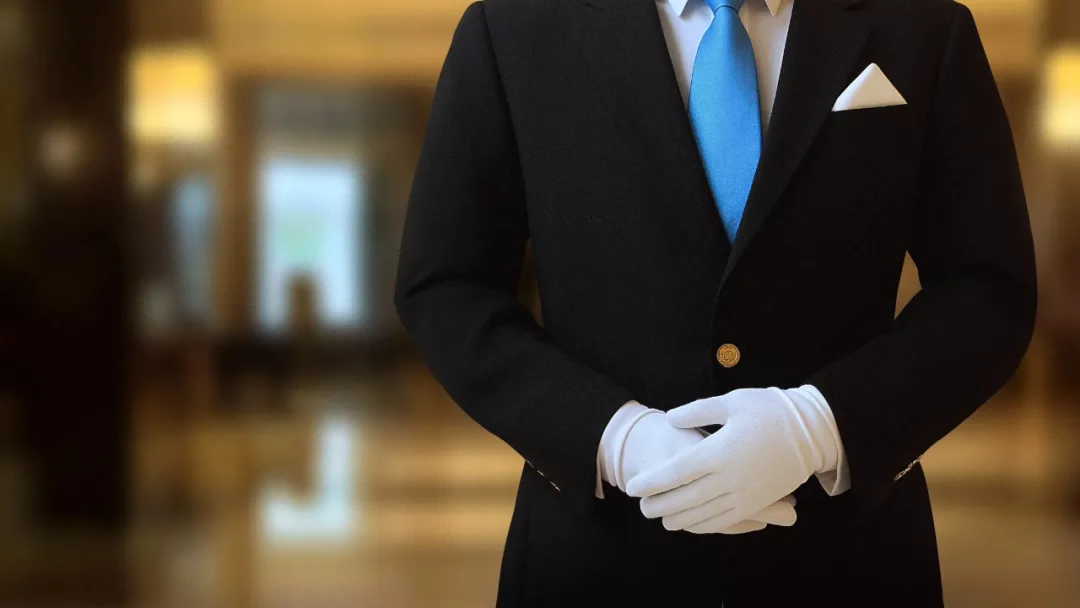 A photo of a professional hotel staff member in a black suit with a blue tie, with their hands politely clasped in front of them in a modern hotel lobby.