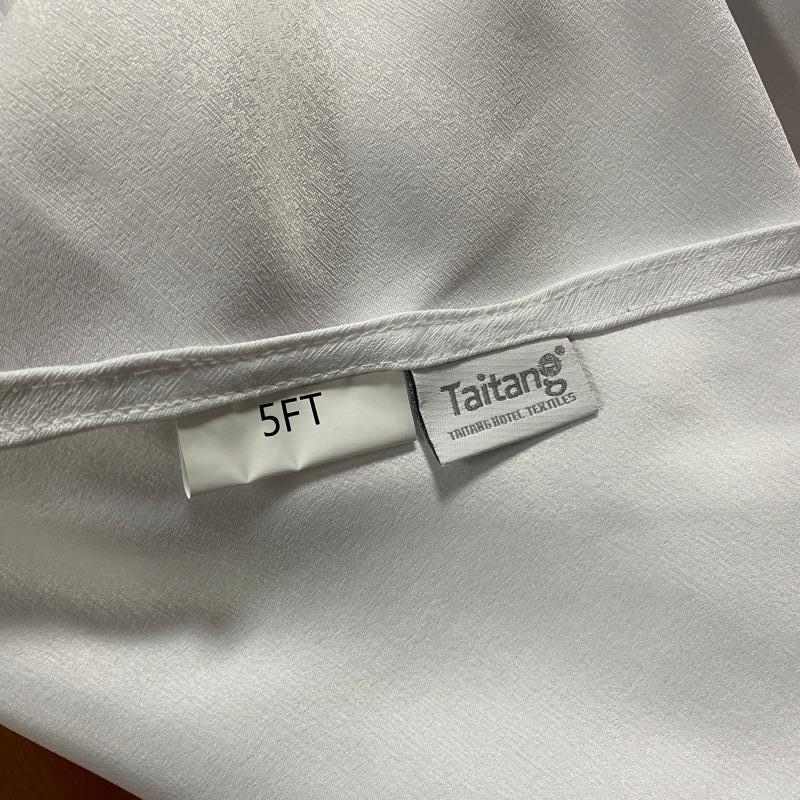 Close-up of white commercial tablecloth hem showing a custom sewn Taitang brand tag and 5FT table size label.JPG