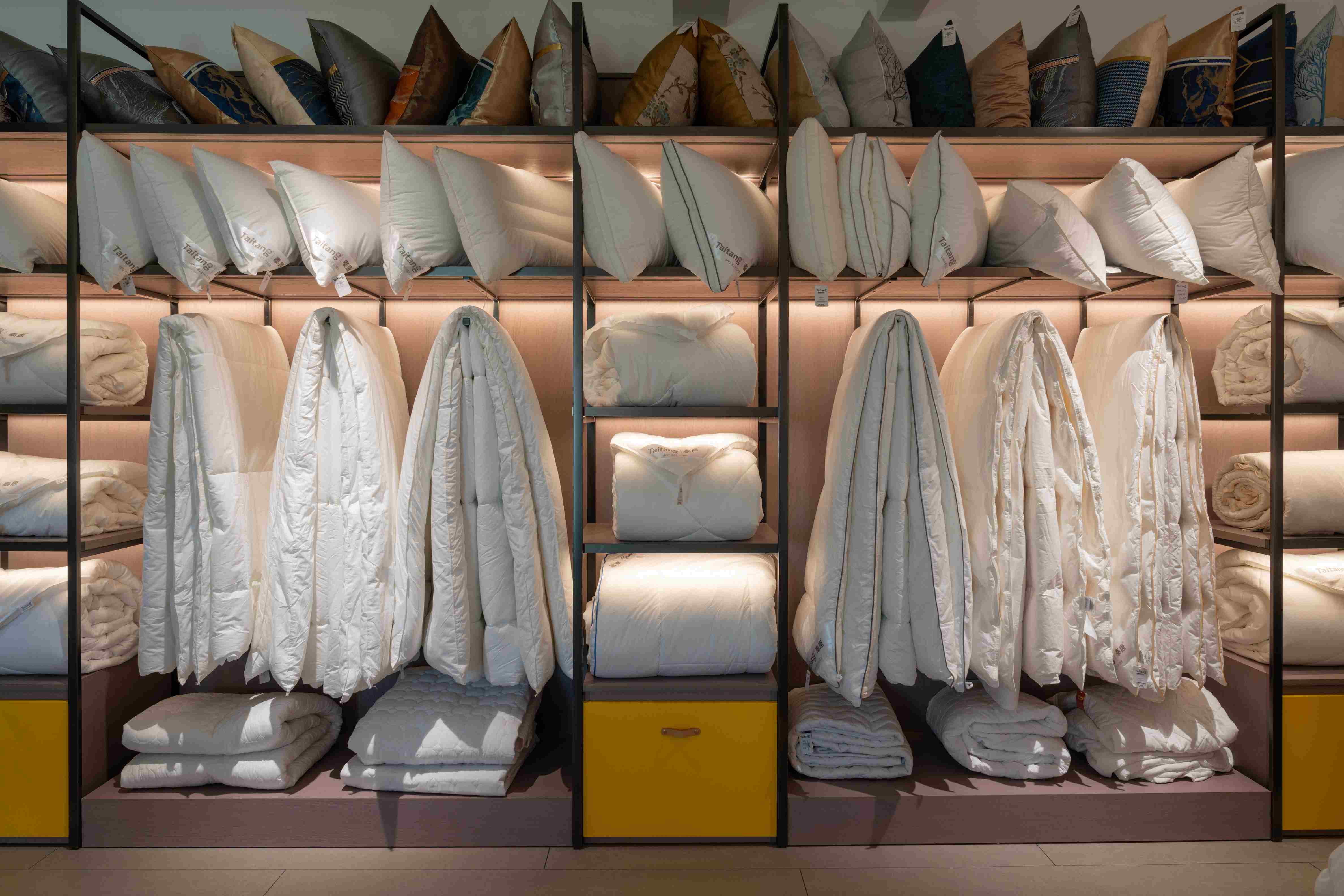 A professional hotel supply showroom display featuring organized shelves of white duvets, colorful pillows, and premium bedding samples.