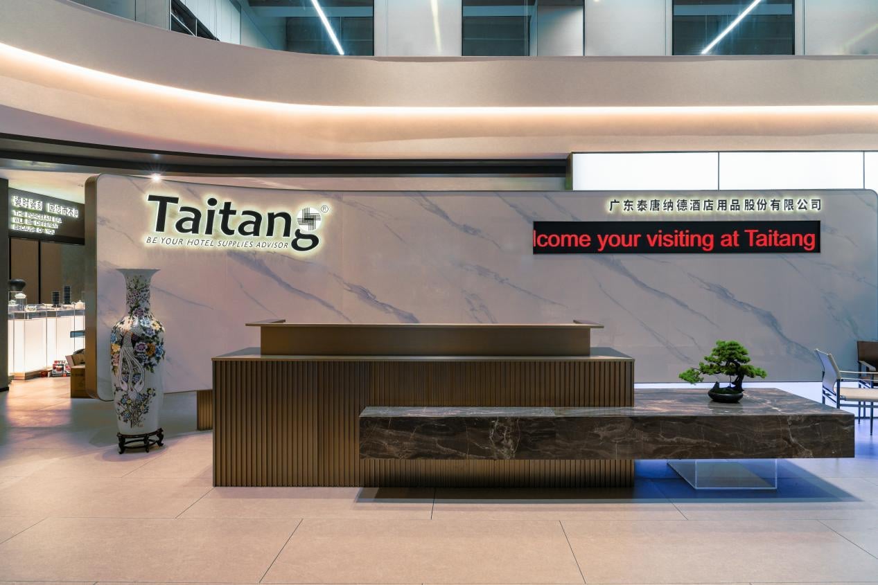 The modern and luxurious reception area of the Taitang showroom, featuring a marble-textured wall with the Taitang logo, a grand decorative vase, and a sleek dark stone front desk.