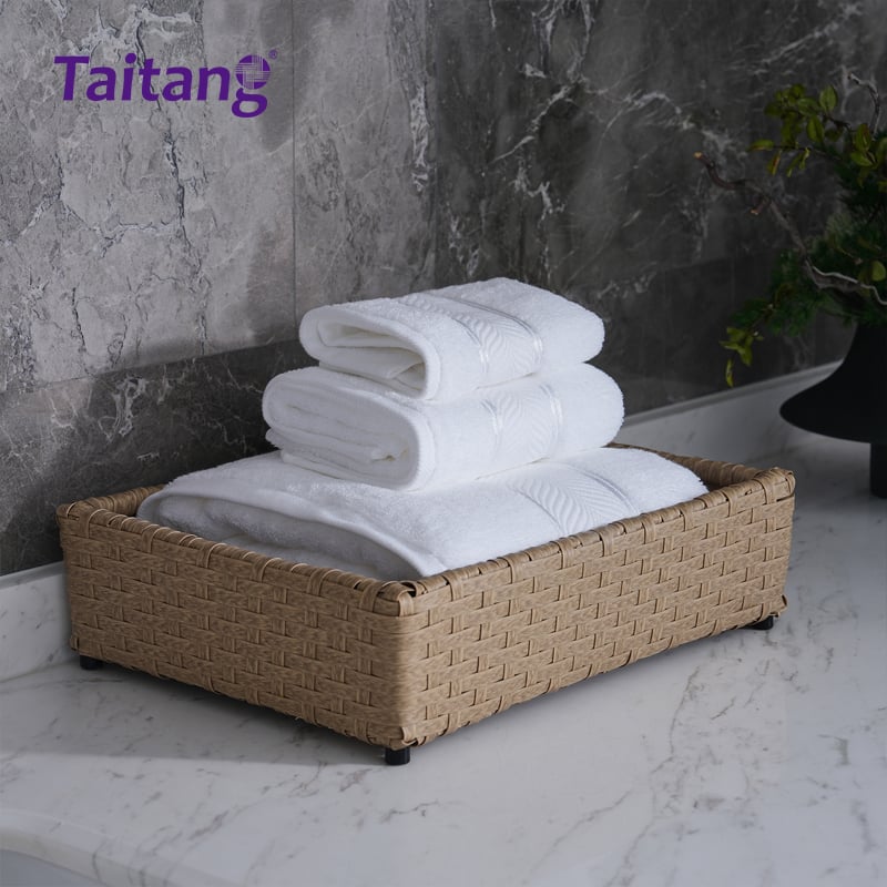 A premium three-piece set of white cotton towels (bath towel, hand towel, and washcloth) neatly folded and stacked in a rectangular woven basket on a white marble countertop.jpg