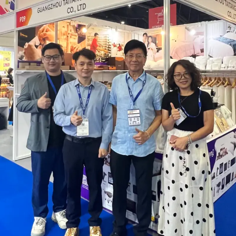 Four smiling colleagues posing in front of their company's booth at a Thai exhibition, giving thumbs-up.webp