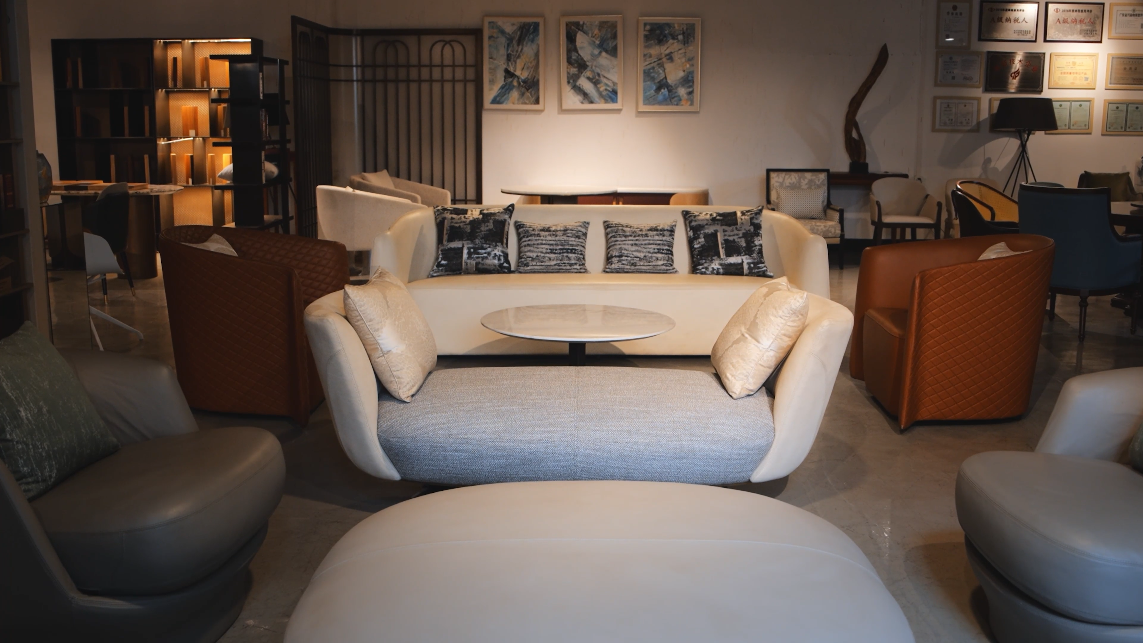 A professional furniture showroom display featuring a modern cream-colored sofa with patterned grey pillows, a circular marble coffee table, and cognac leather armchairs in a well-lit, sophisticated lounge setting.