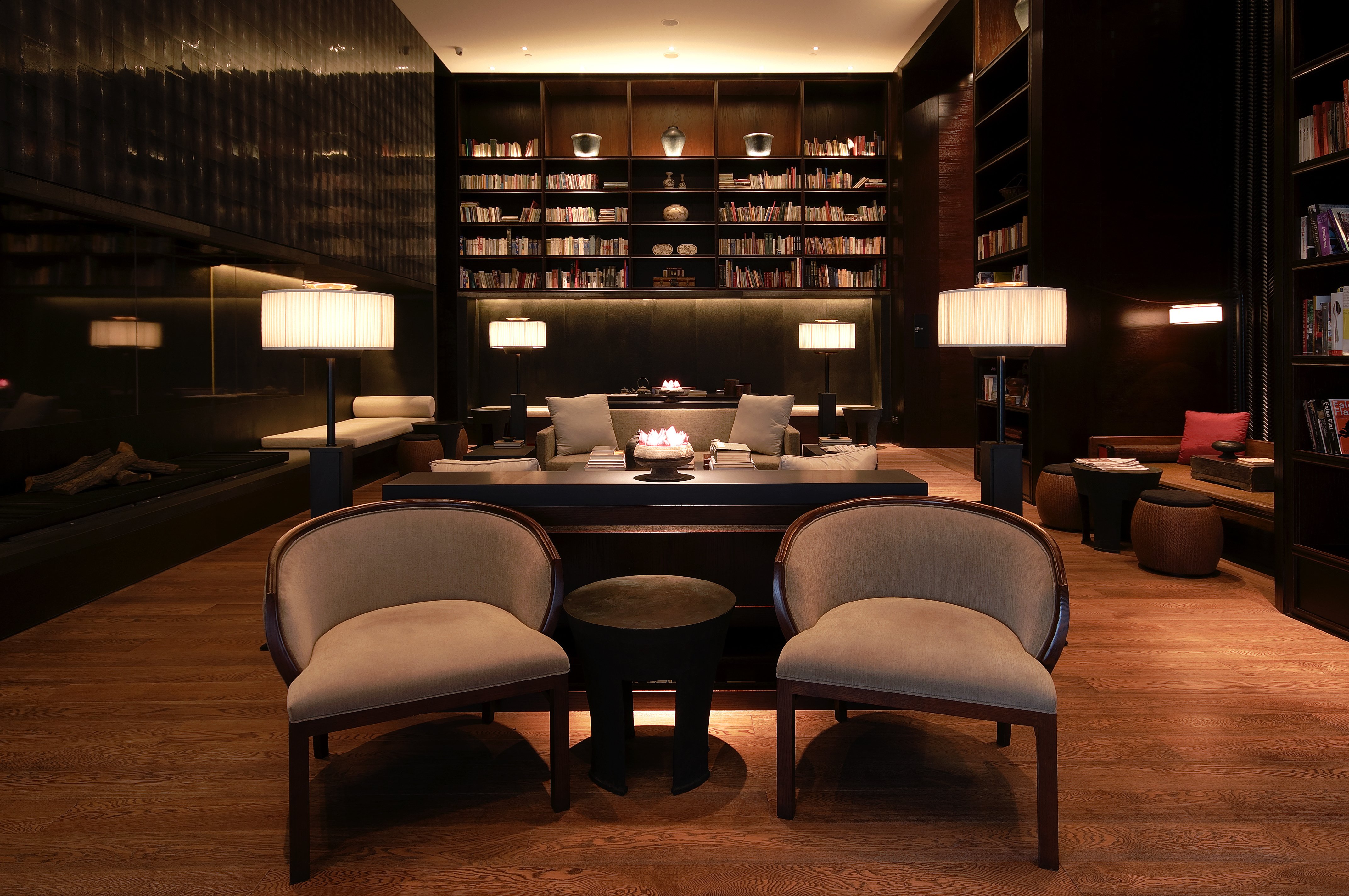 A sophisticated hotel library and lounge area with warm ambient lighting, featuring floor-to-ceiling dark wood bookshelves, elegant upholstered armchairs, and minimalist floor lamps in a cozy, upscale atmosphere.
