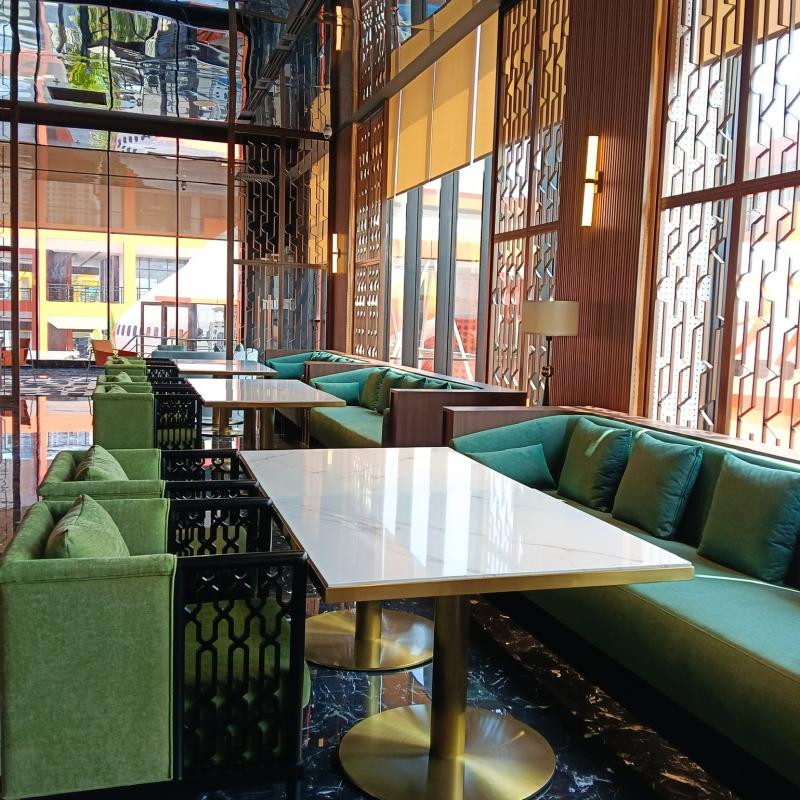 Bright hotel dining lounge with emerald green velvet booth seating and marble-top restaurant tables.webp