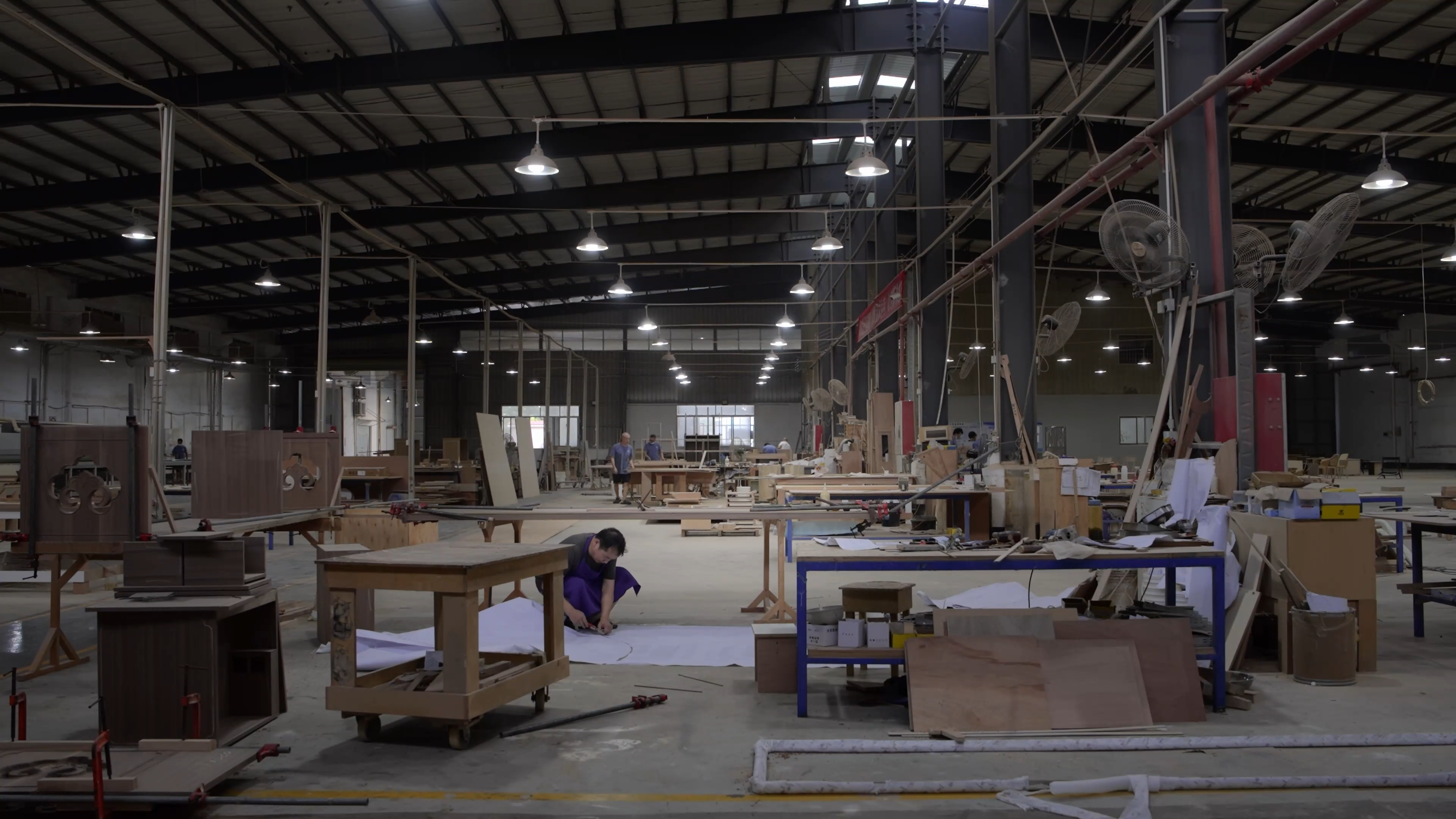 A wide-angle view of a large furniture manufacturing factory and workshop, showing workers assembling wooden furniture frames and components in a spacious industrial facility with high ceilings and metal beams.