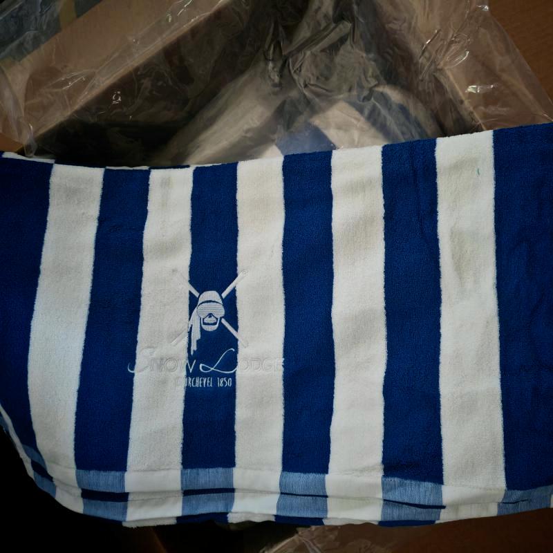 A top-down view looking into an opened brown cardboard box branded with the Taitang logo. Inside, a blue and white striped bath or spa towel is folded.jpg