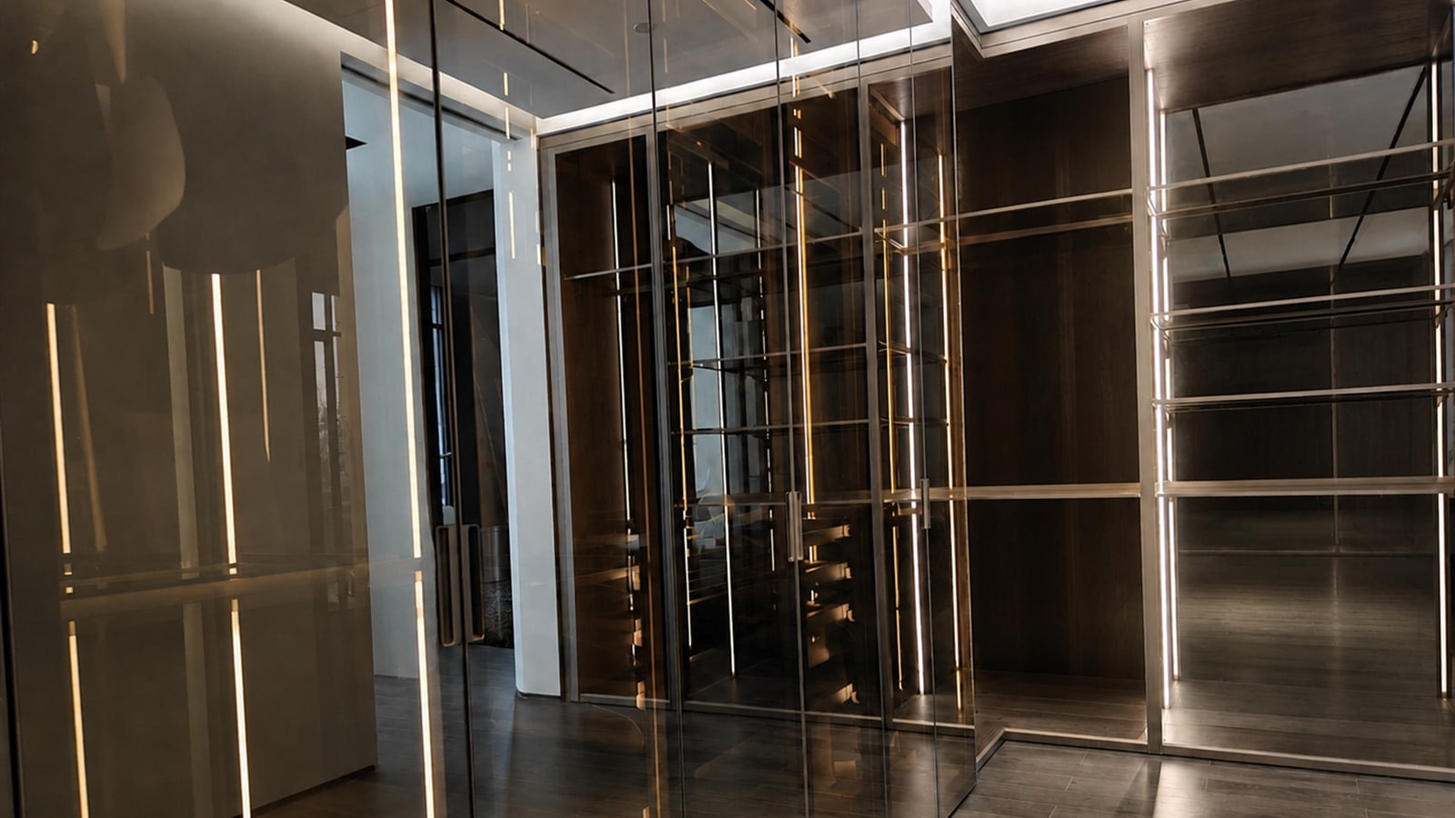 Completed floor-to-ceiling glass wardrobe installation with slim aluminum frames, wood-finish storage and integrated lighting