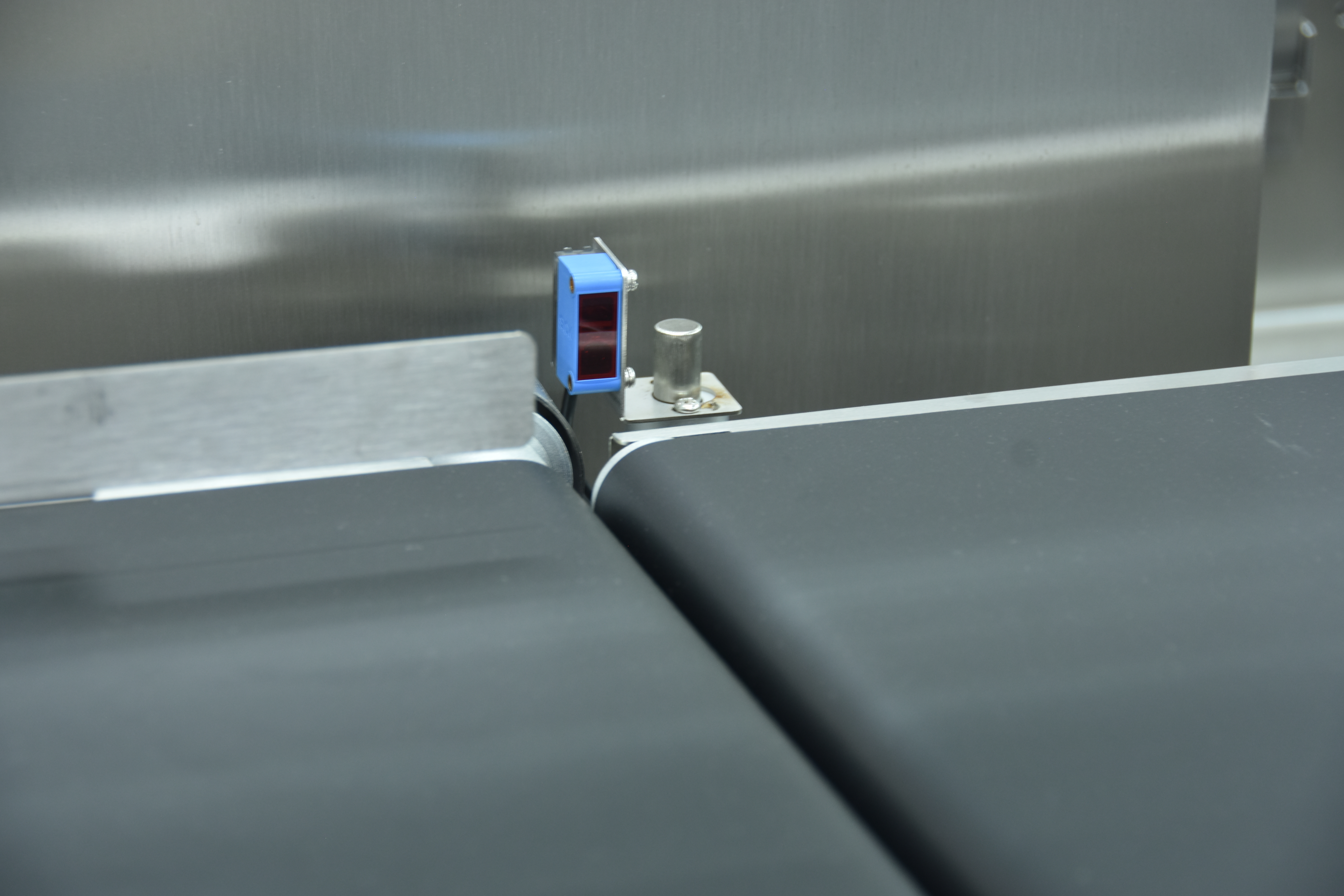 Close-up of the sensor at the transfer between two belt sections: a blue rectangular photoelectric sensor with a dark window, bolted to a stainless angle bracket, standing beside a stainless cylindrical stud on a plate, with brushed stainless panelling behind and the dark belt and its roller edge below