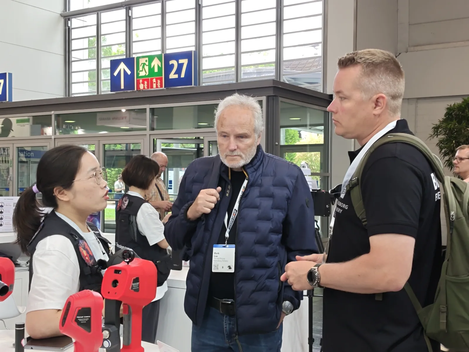 IMG-03 | Handheld Thermal Imager Demonstration LSJ specialist explaining a red handheld thermal imaging camera to two international visitors at the Hannover Fire and Rescue Exhibition