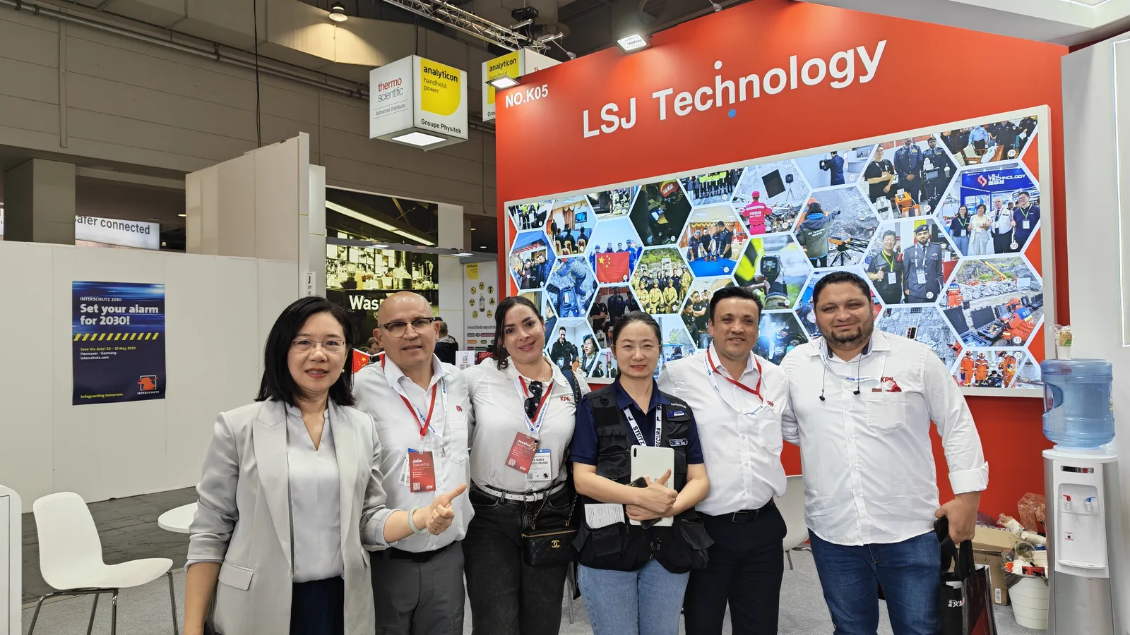 IMG-07 | LSJ Team with International Client Delegation LSJ team members posing with an international client delegation in front of the red LSJ Technology photo wall at booth NO.K05 in Hannover